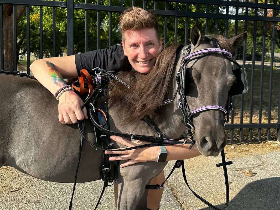 The author, an animal communicator, hugging their miniature horse Sabi, who is wearing a black harness with purple trim.